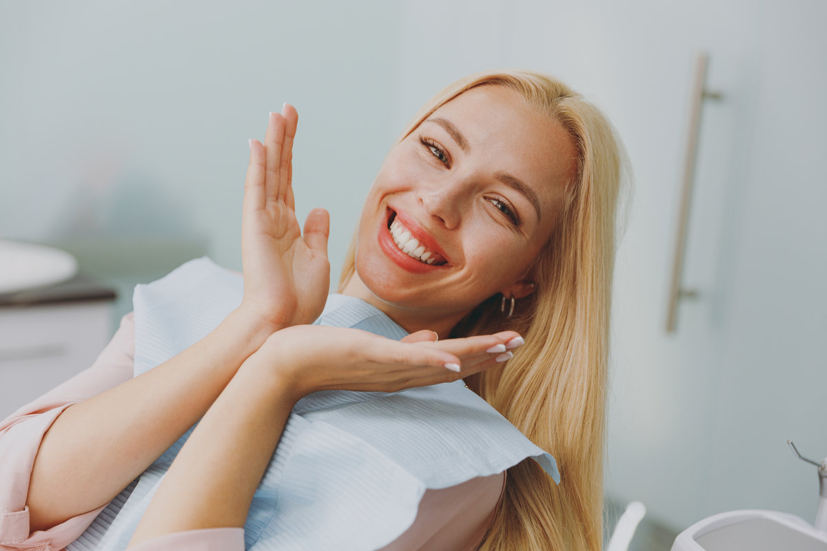 Close up young amazed smiling happy woman covered by napkin spread hands sit at dental office chair indoor light modern cabinet wait stomatologist oral procedure. Healthcare caries enamel treatment.